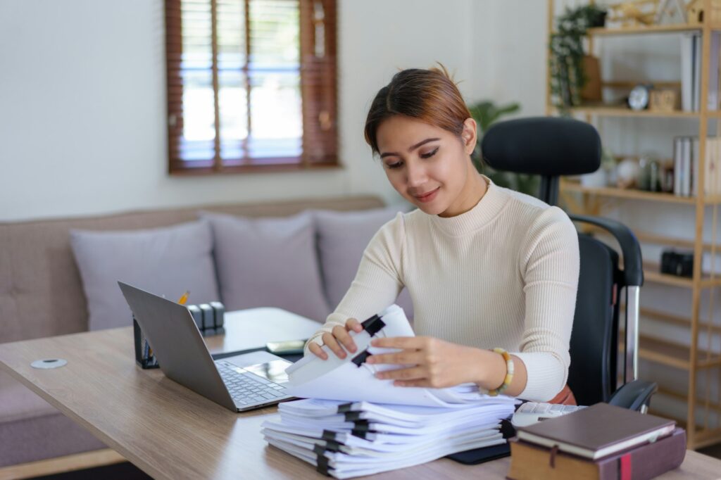Focused asian woman organizing paperwork at a home office desk with a laptop.